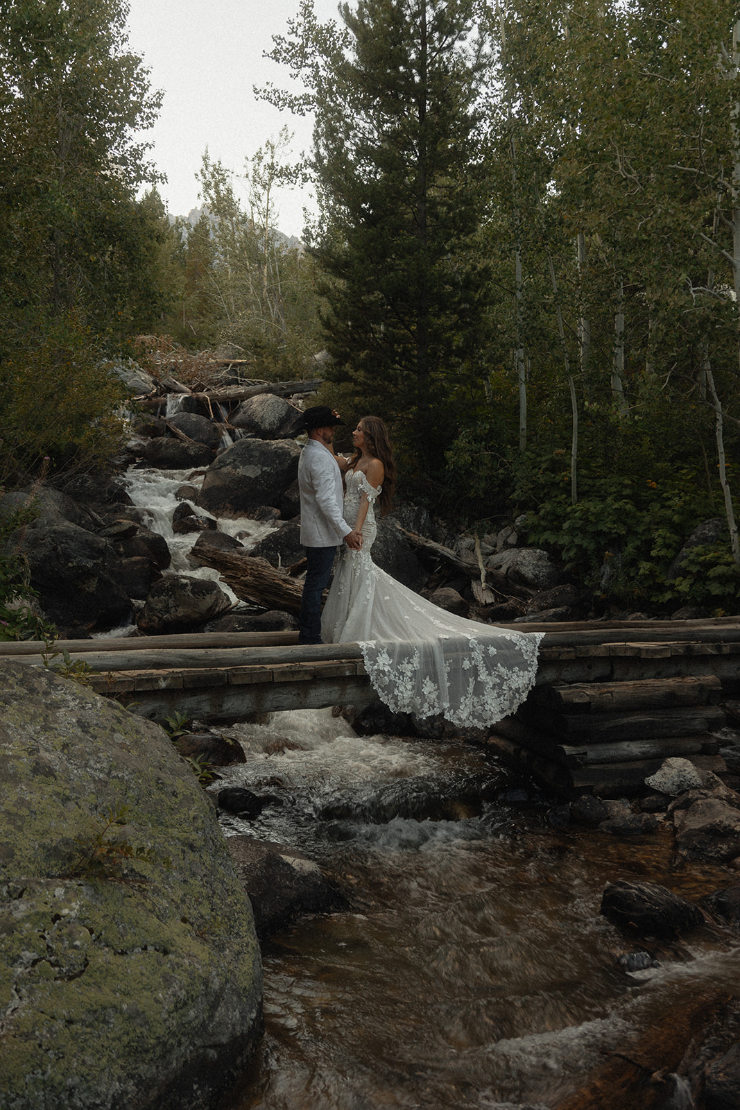 A couple stands on a wooden bridge in a forested area. The woman wears a long, white lace gown while the man wears a white jacket and dark pants. Trees and a stream are visible in the background for a Grand Teton National park taggart lake elopement