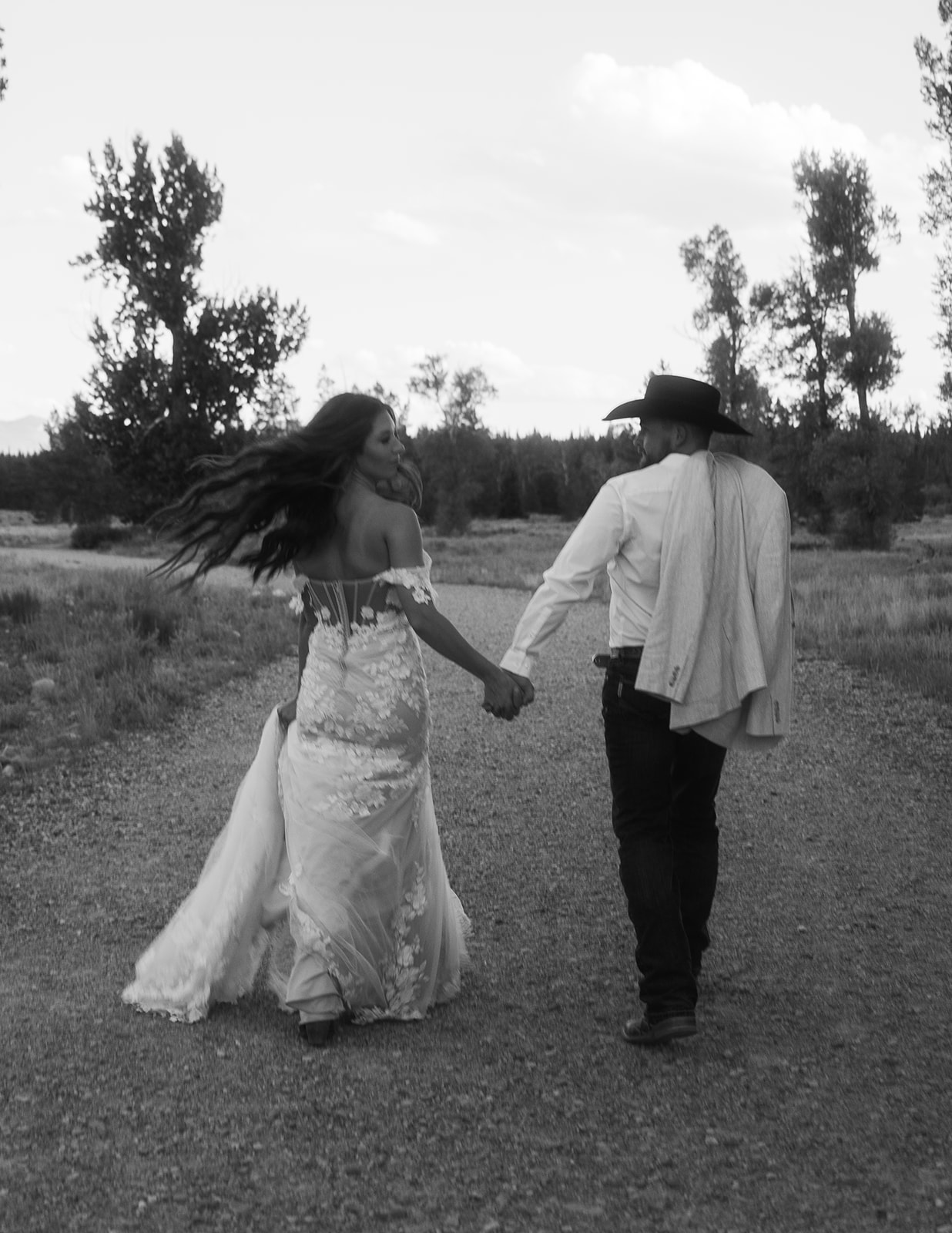 A couple stands on a wooden bridge in a forested area. The woman wears a long, white lace gown while the man wears a white jacket and dark pants. Trees and a stream are visible in the background for a Grand Teton National park taggart lake elopement