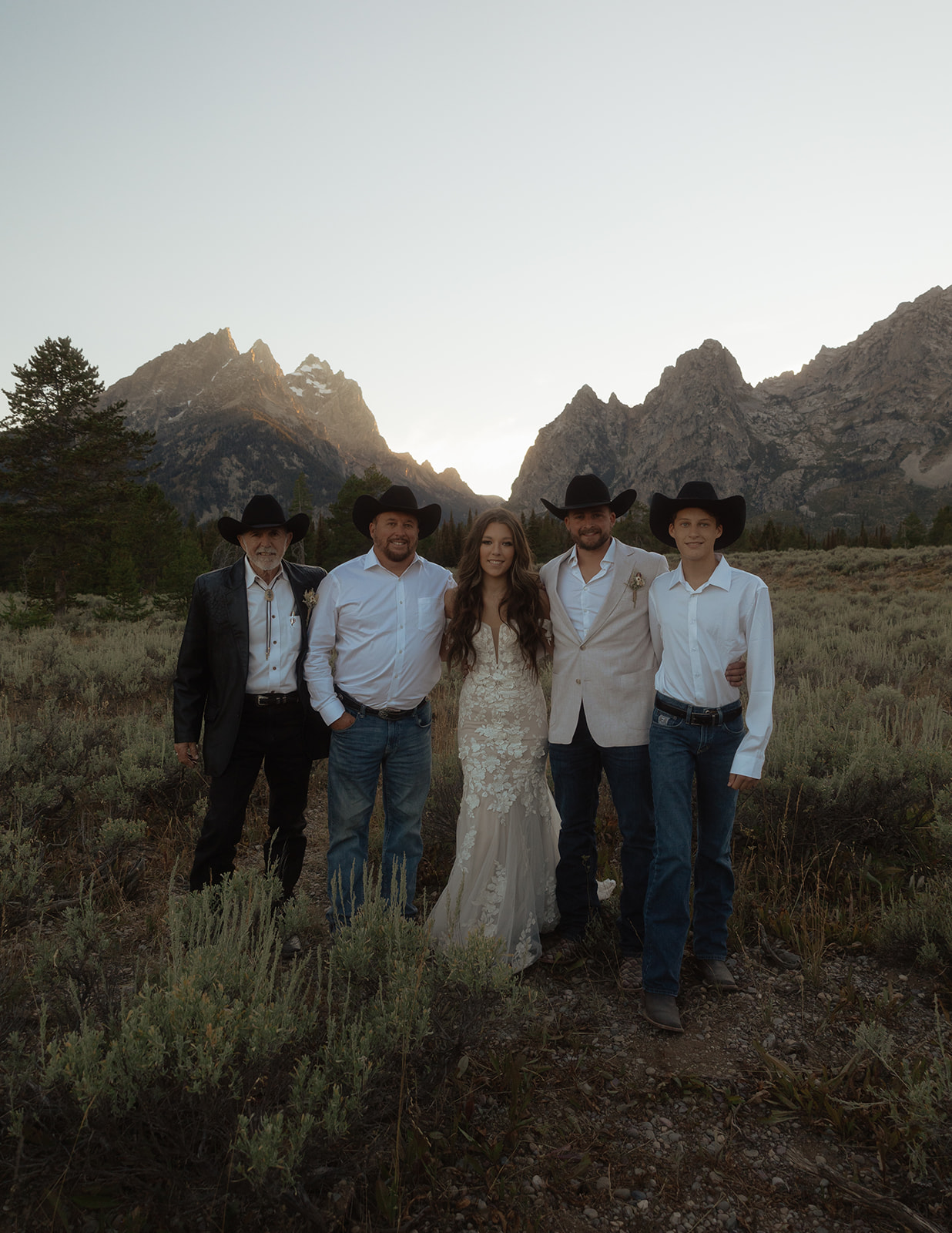 Five people, dressed in Western attire and cowboy hats, stand together in a mountainous landscape at sunset at Grand Teton National park taggart lake