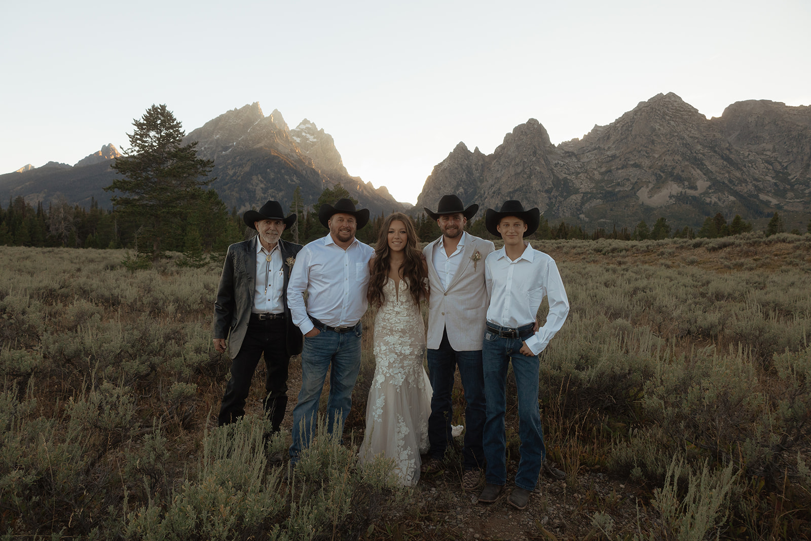 Five people, dressed in Western attire and cowboy hats, stand together in a mountainous landscape at sunset at Grand Teton National park taggart lake