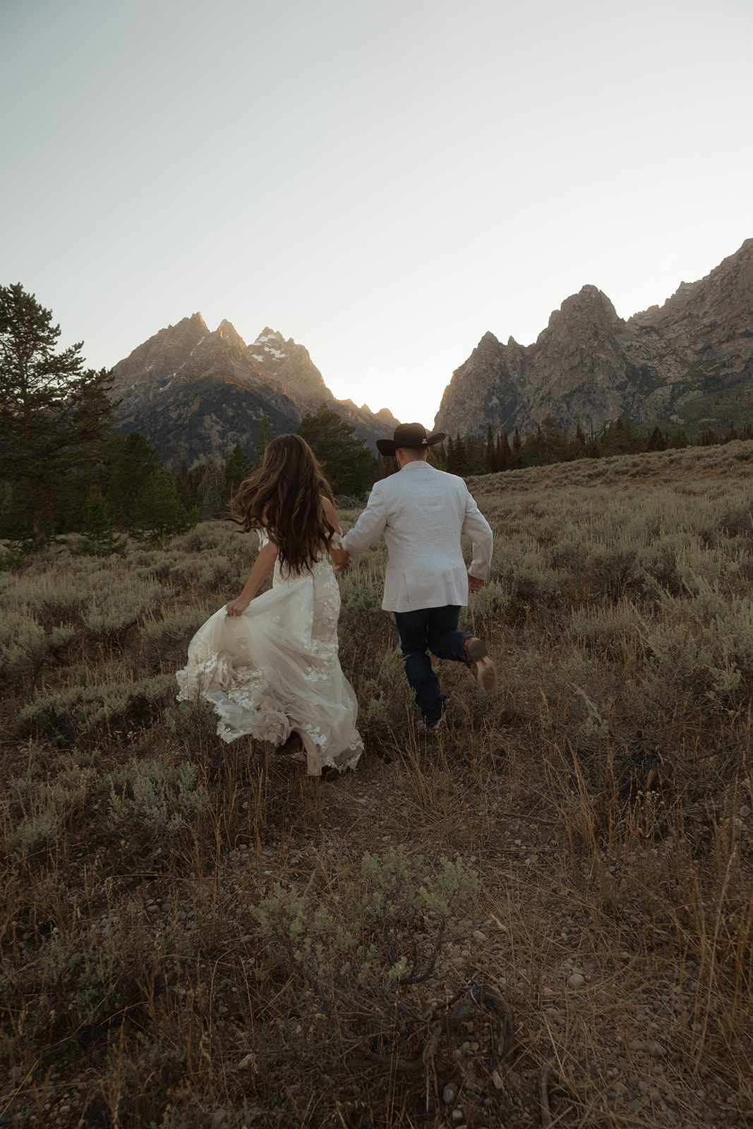 A couple in wedding attire, with the woman in a white dress and the man in a white shirt and black hat, walking hand in hand through a field toward mountains at sunset at Grand Teton National park taggart lake