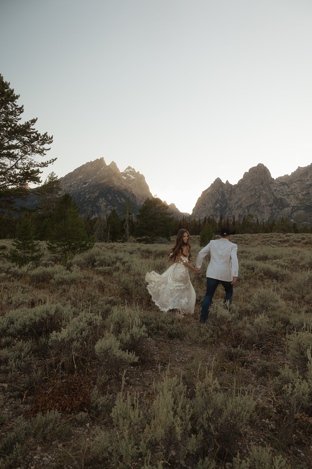 A couple in wedding attire, with the woman in a white dress and the man in a white shirt and black hat, walking hand in hand through a field toward mountains at sunset at taggart lake