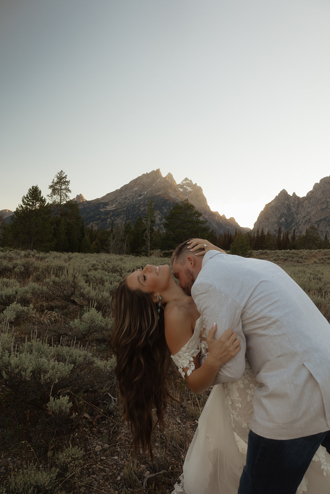 A couple embraces with mountains in the background at sunset, the woman wearing a white dress and the man in a light-colored shirt at Grand Teton National park Taggart lake for an elopement