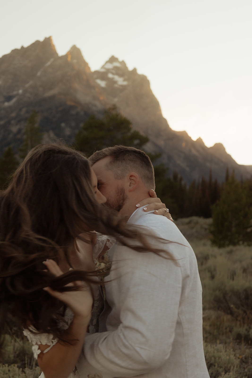 A couple embraces with mountains in the background at sunset, the woman wearing a white dress and the man in a light-colored shirt at Taggart lake for an elopement