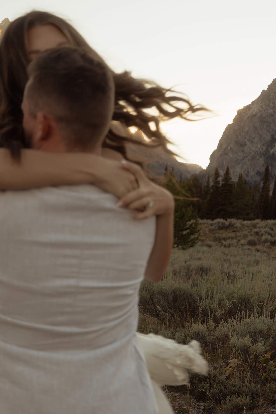 A couple embraces with mountains in the background at sunset, the woman wearing a white dress and the man in a light-colored shirt at Taggart lake for an elopement