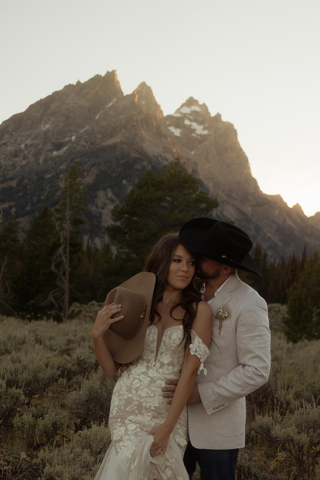 A couple embraces with mountains in the background at sunset, the woman wearing a white dress and the man in a light-colored shirt at Taggart lake for an elopement