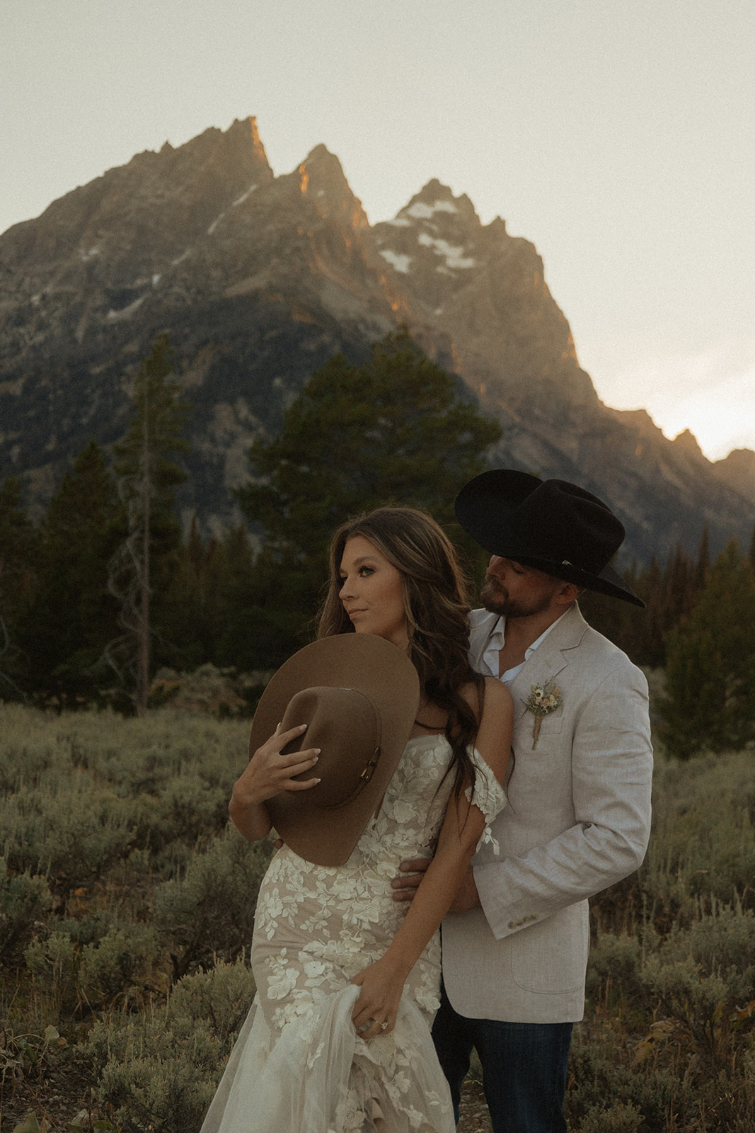 A couple embraces with mountains in the background at sunset, the woman wearing a white dress and the man in a light-colored shirt at Taggart lake for an elopement