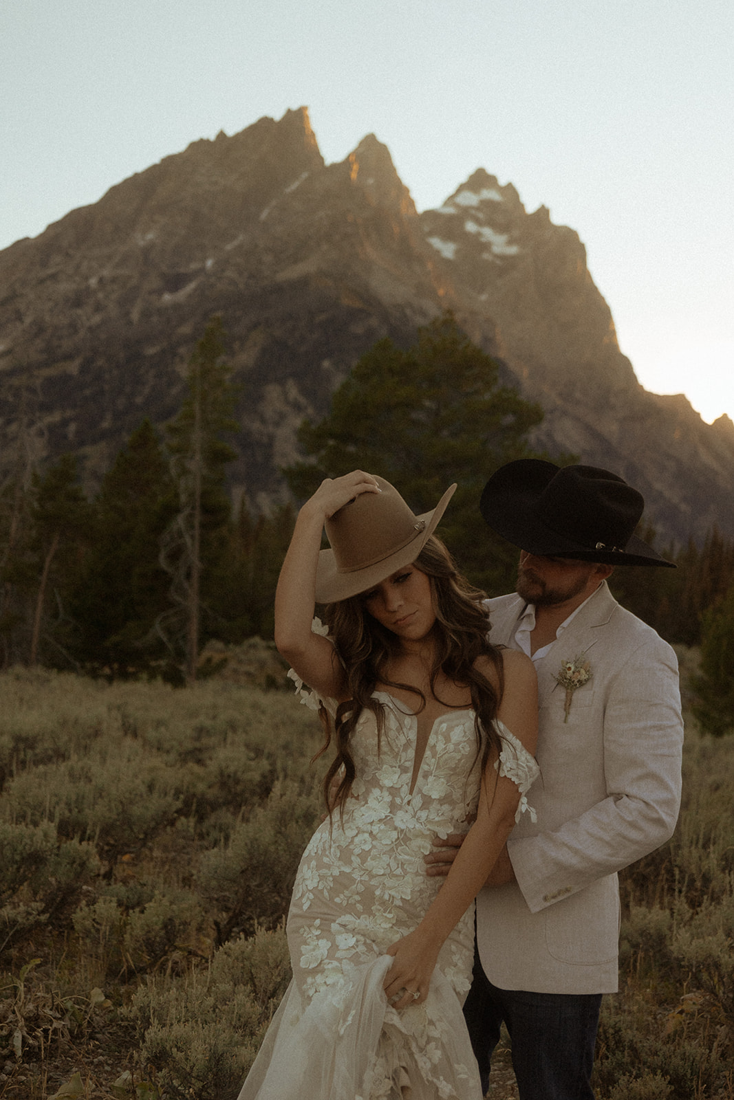 A couple dressed in wedding attire, with cowboy hats, stands embracing in front of a mountainous landscape.