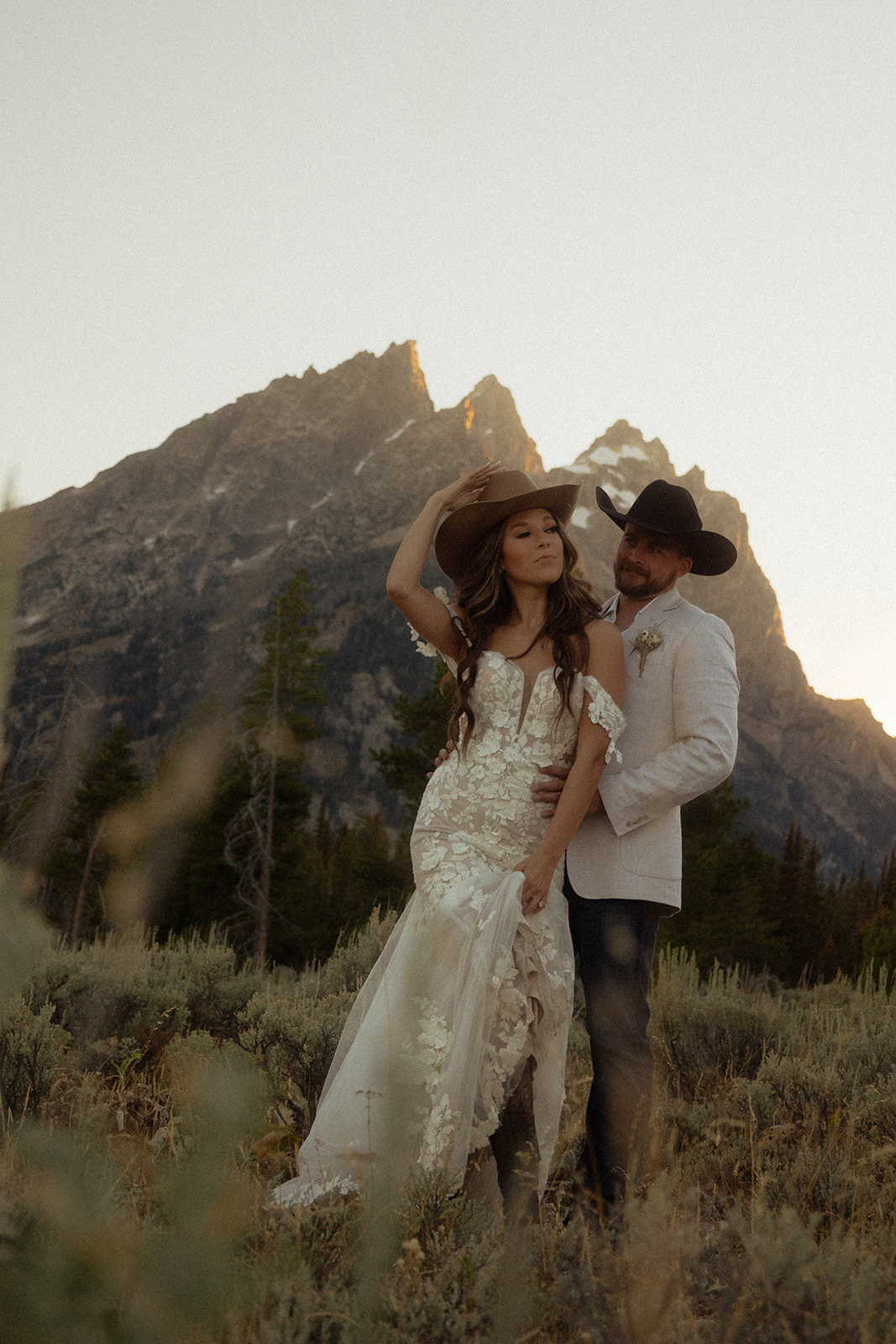 A couple dressed in wedding attire, with cowboy hats, stands embracing in front of a mountainous landscape.