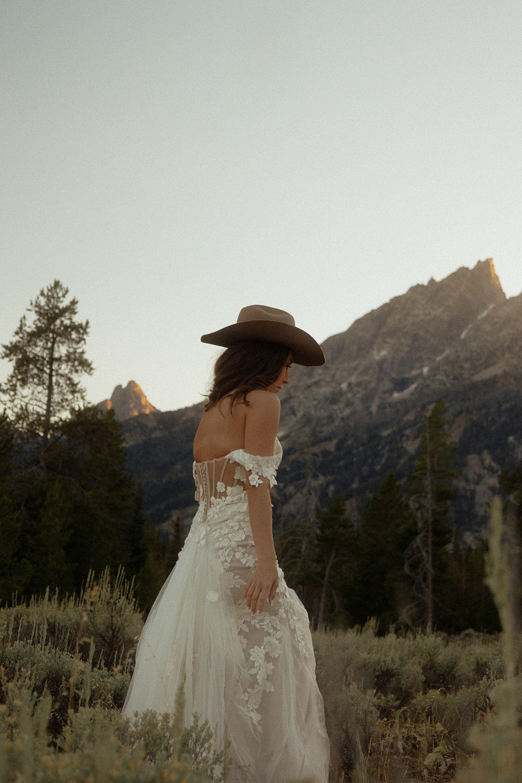 A person in a white dress and wide-brimmed hat stands in a natural landscape with mountains and trees in the background.