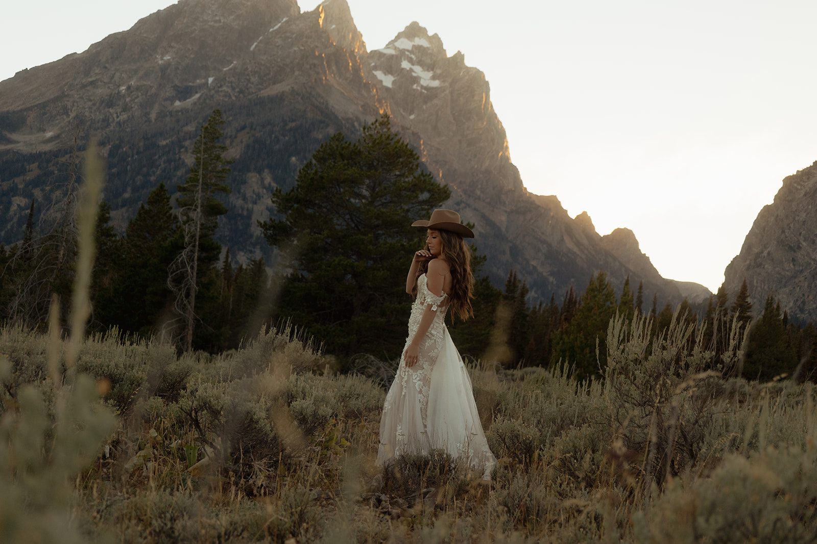 A person in a white dress and wide-brimmed hat stands in a natural landscape with mountains and trees in the background.