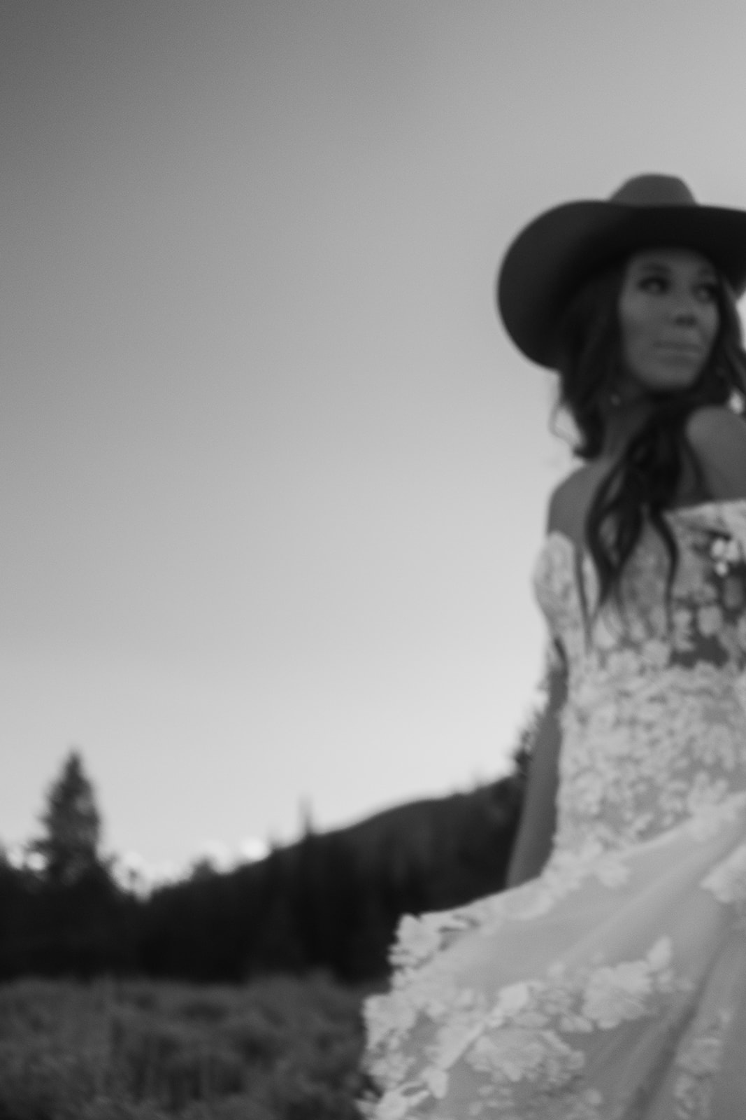 A person in a white dress and wide-brimmed hat stands in a natural landscape with mountains and trees in the background.