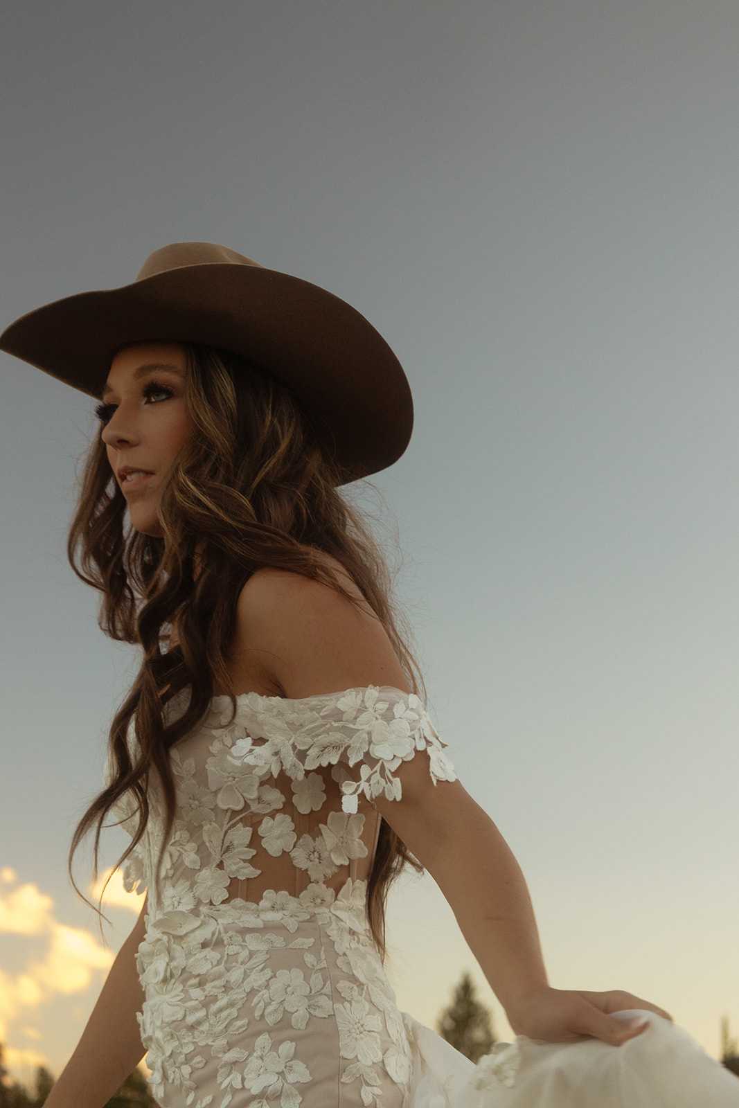 A person in a white dress and wide-brimmed hat stands in a natural landscape with mountains and trees in the background.