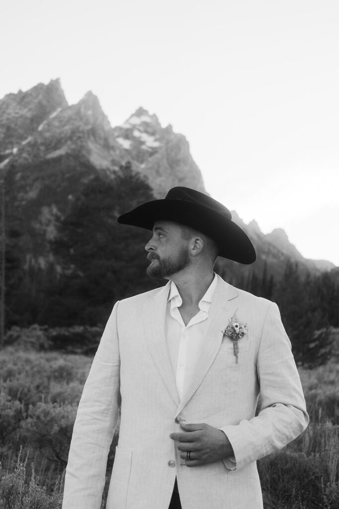 A man in a white blazer and black cowboy hat stands in a mountainous landscape with shrubs and trees.