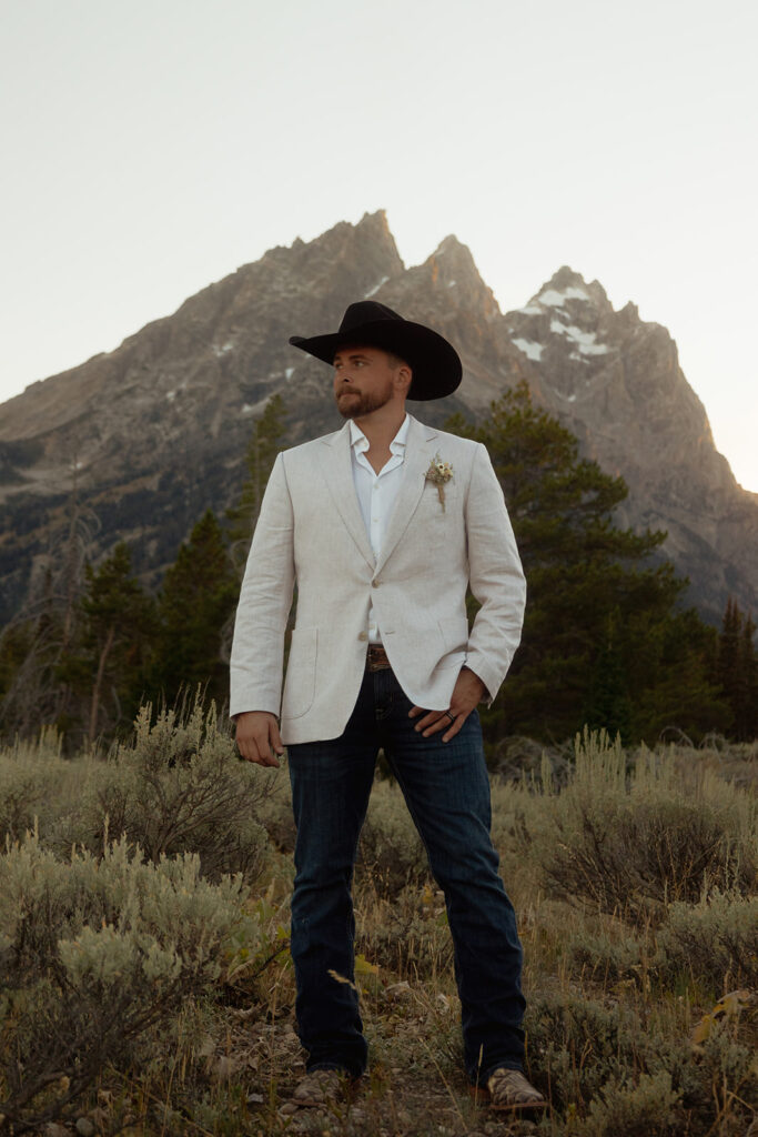 A man in a white blazer and black cowboy hat stands in a mountainous landscape with shrubs and trees.