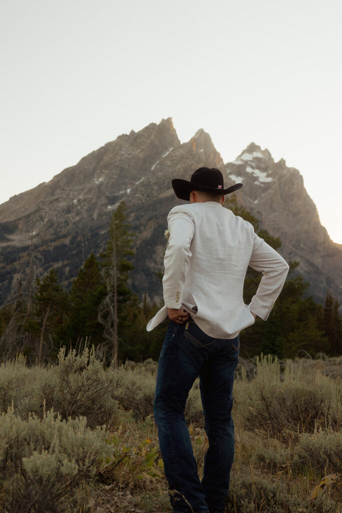 A man in a white blazer and black cowboy hat stands in a mountainous landscape with shrubs and trees.