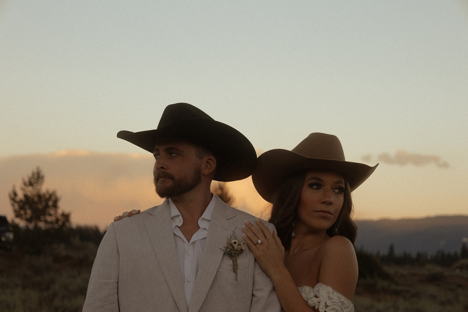 A couple wearing formal western attire pose outdoors at sunset. Both have cowboy hats; the man wears a white blazer with a boutonniere and the woman wears an off-shoulder dress.