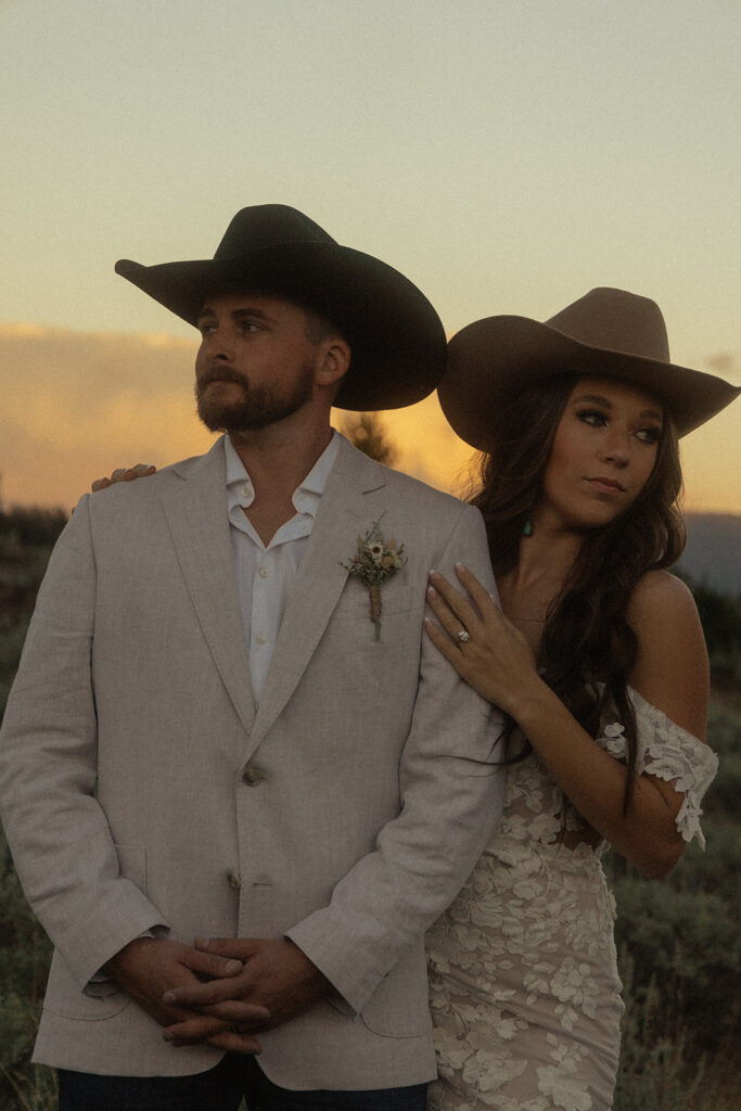 A couple wearing formal western attire pose outdoors at sunset. Both have cowboy hats; the man wears a white blazer with a boutonniere and the woman wears an off-shoulder dress.