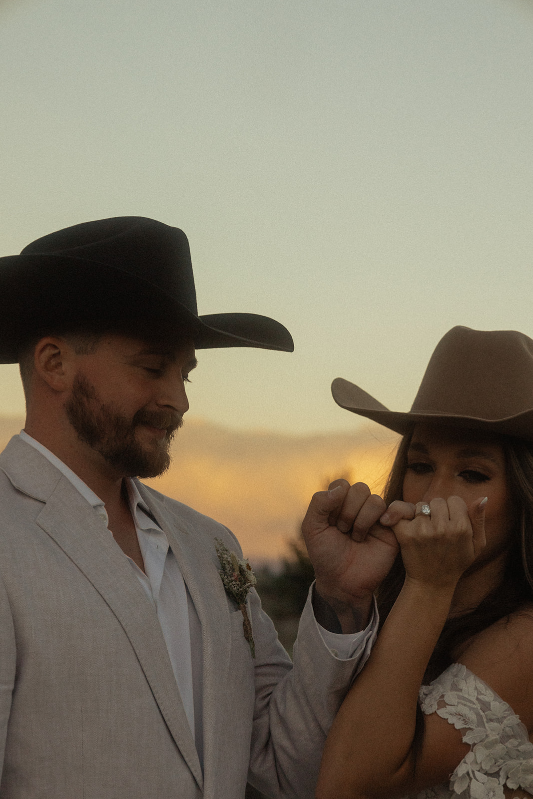 A couple wearing formal western attire pose outdoors at sunset. Both have cowboy hats; the man wears a white blazer with a boutonniere and the woman wears an off-shoulder dress.