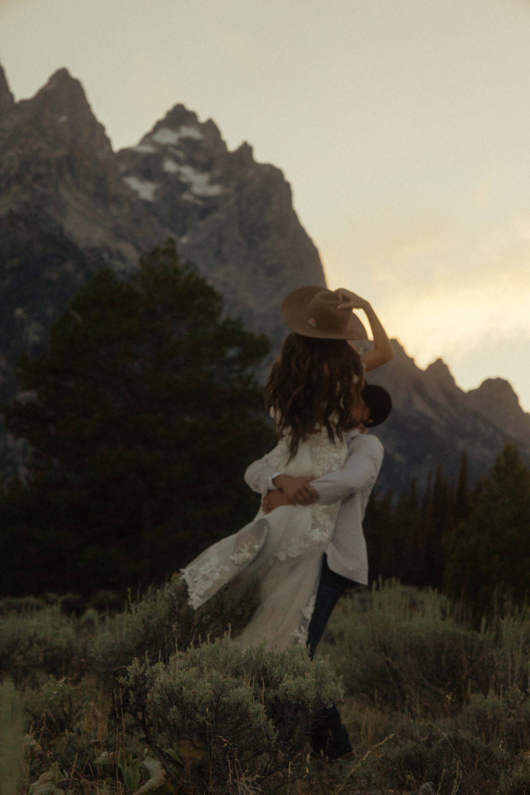 A person lifts and embraces another in a field with tall mountain peaks and trees in the background.