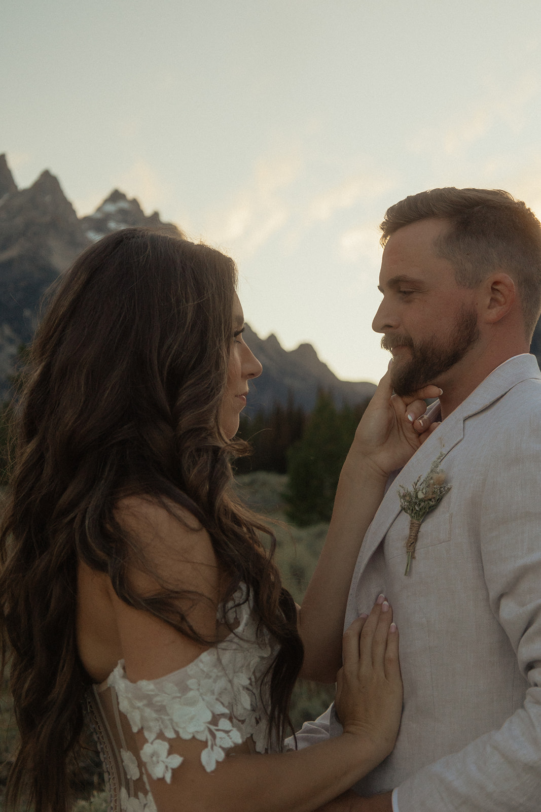 A person lifts and embraces another in a field with tall mountain peaks and trees in the background.