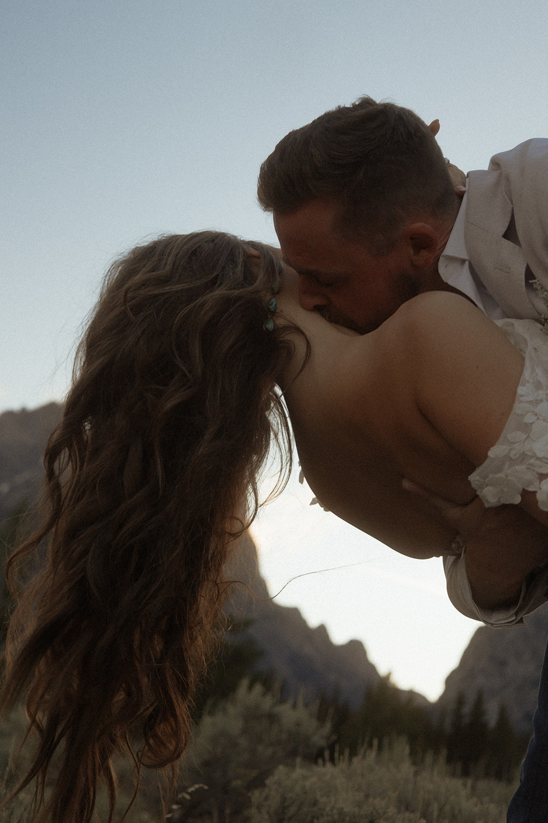 A person lifts and embraces another in a field with tall mountain peaks and trees in the background.