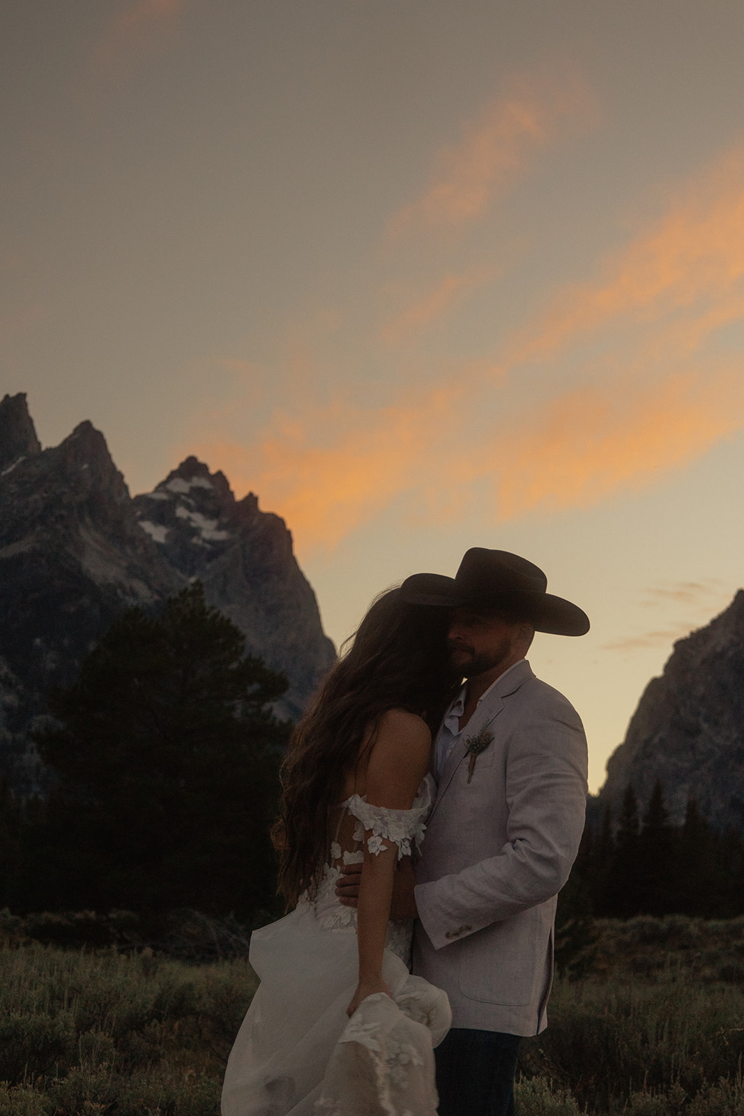 A person lifts and embraces another in a field with tall mountain peaks and trees in the background.