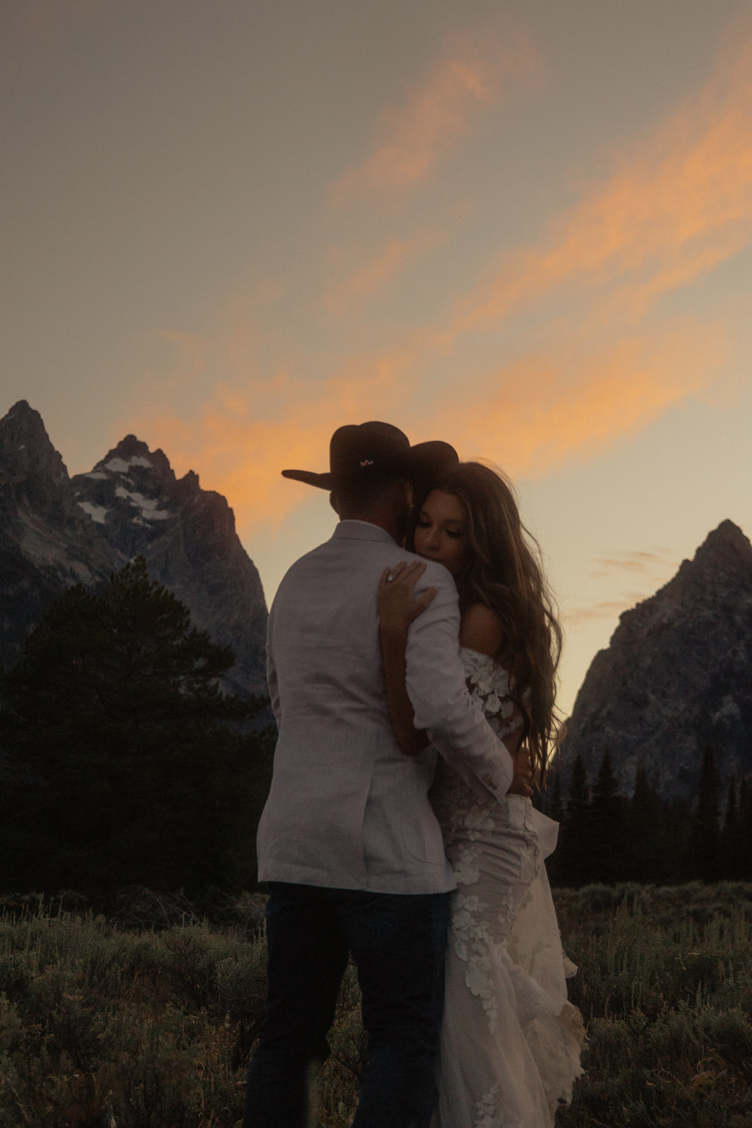 A person lifts and embraces another in a field with tall mountain peaks and trees in the background.