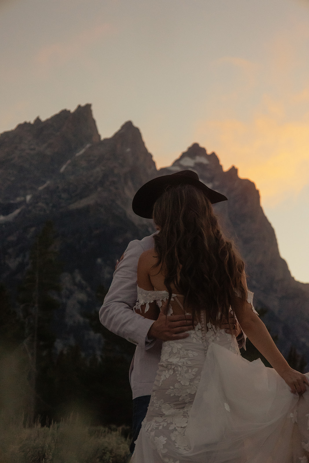 A person lifts and embraces another in a field with tall mountain peaks and trees in the background.