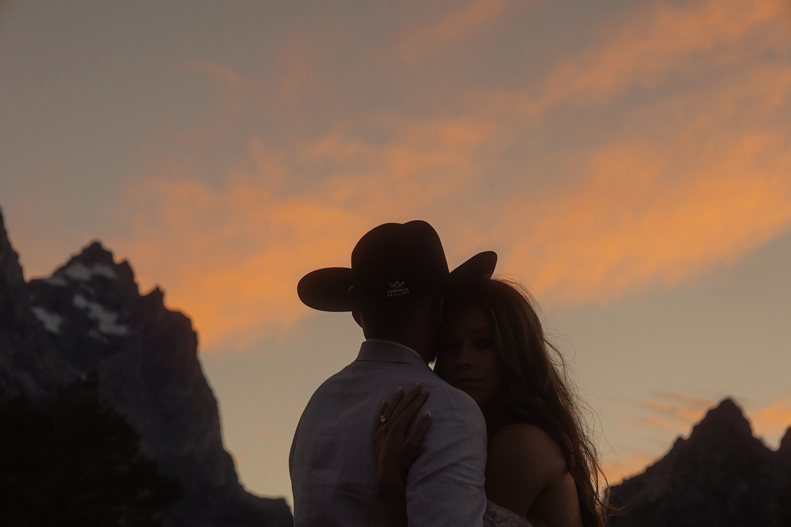 A person lifts and embraces another in a field with tall mountain peaks and trees in the background.