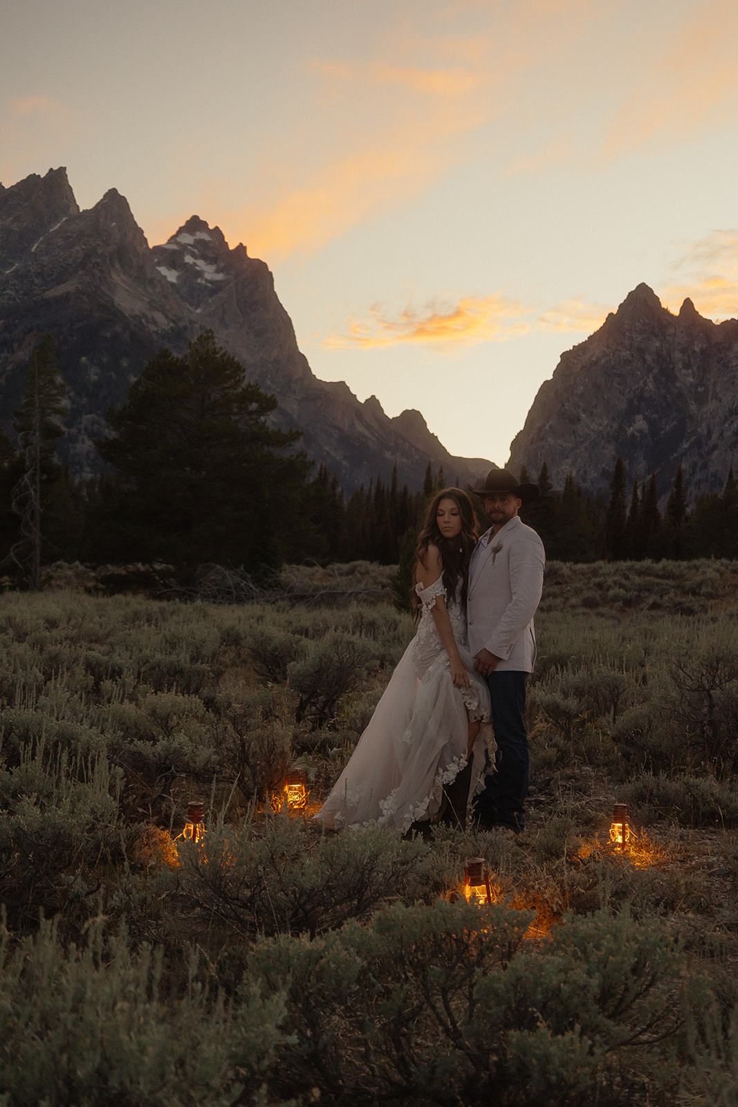 A person lifts and embraces another in a field with tall mountain peaks and trees in the background.