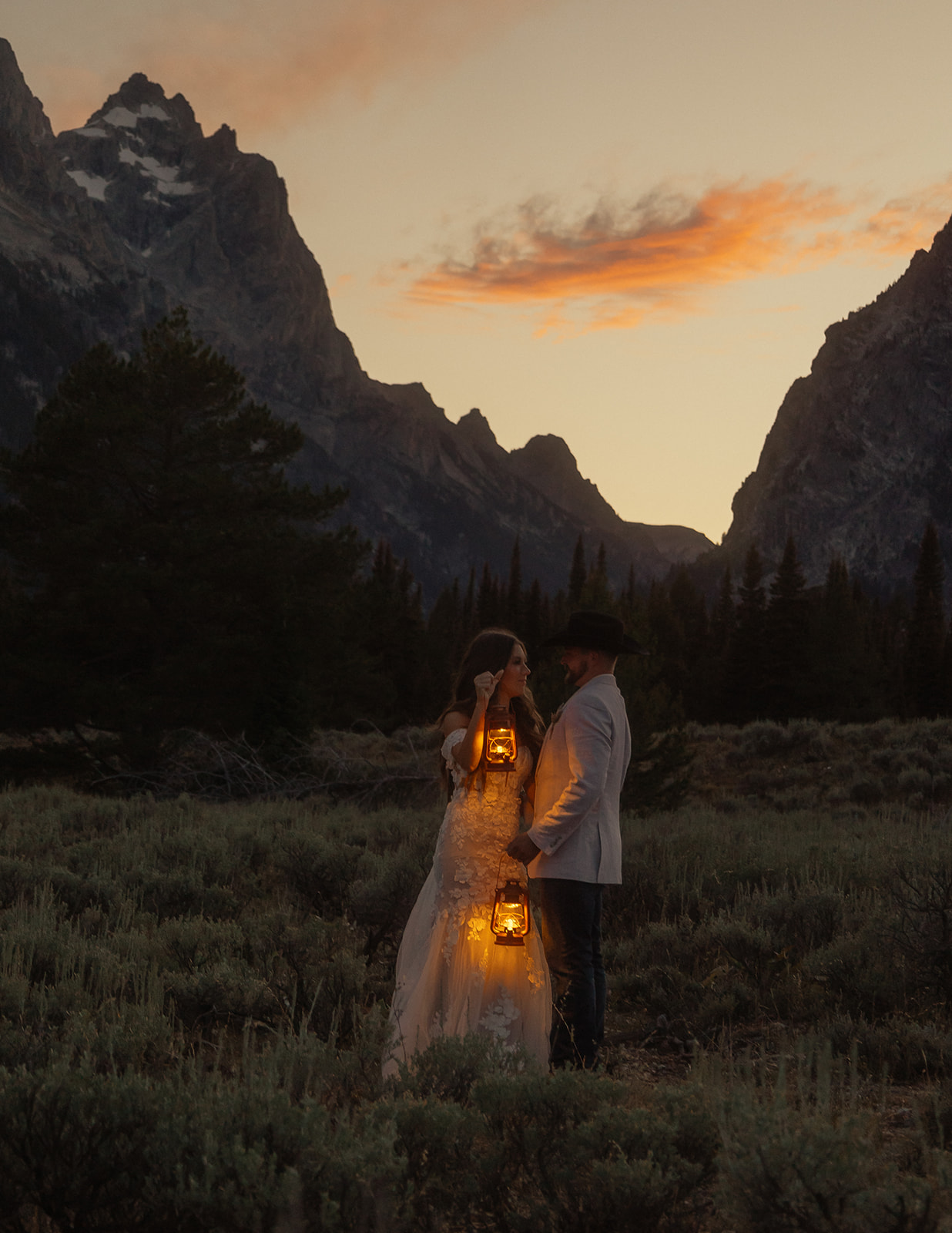 A person lifts and embraces another in a field with tall mountain peaks and trees in the background.