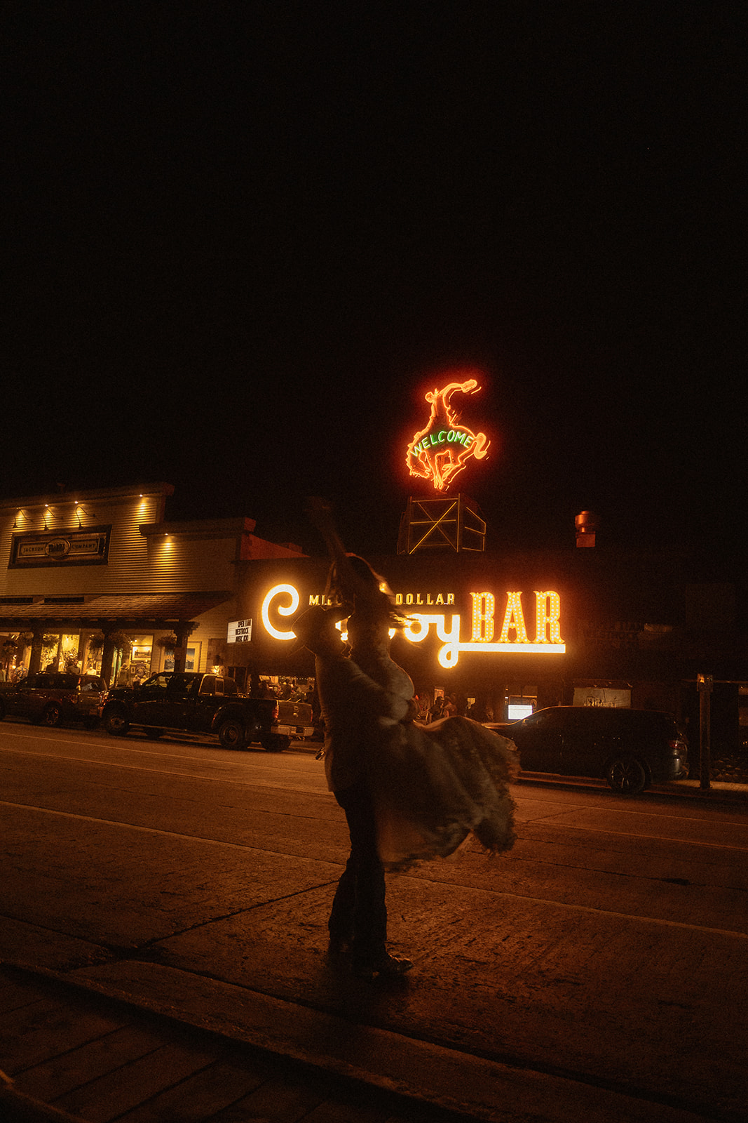 Person standing on a road at night, illuminated by a neon sign for the Cowboy Bar. Nearby building lights are also visible in the background.