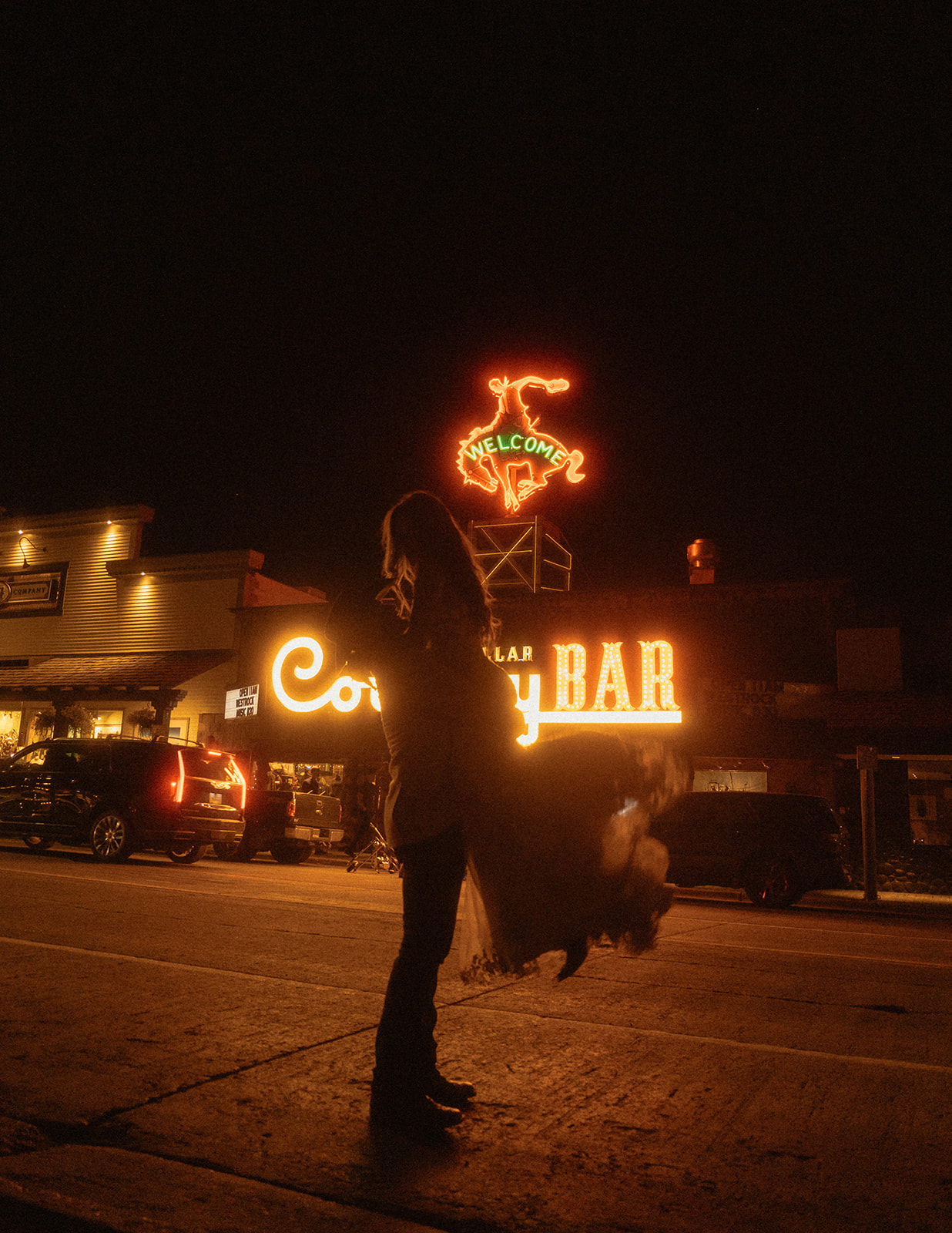 Person standing on a road at night, illuminated by a neon sign for the Cowboy Bar. Nearby building lights are also visible in the background.