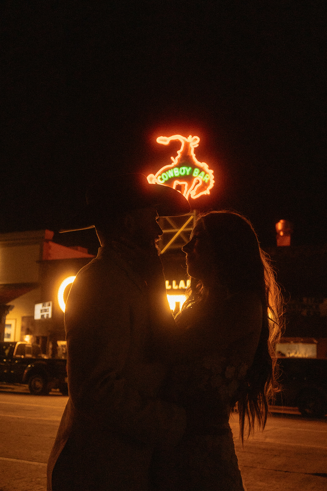 Person standing on a road at night, illuminated by a neon sign for the Cowboy Bar. Nearby building lights are also visible in the background.