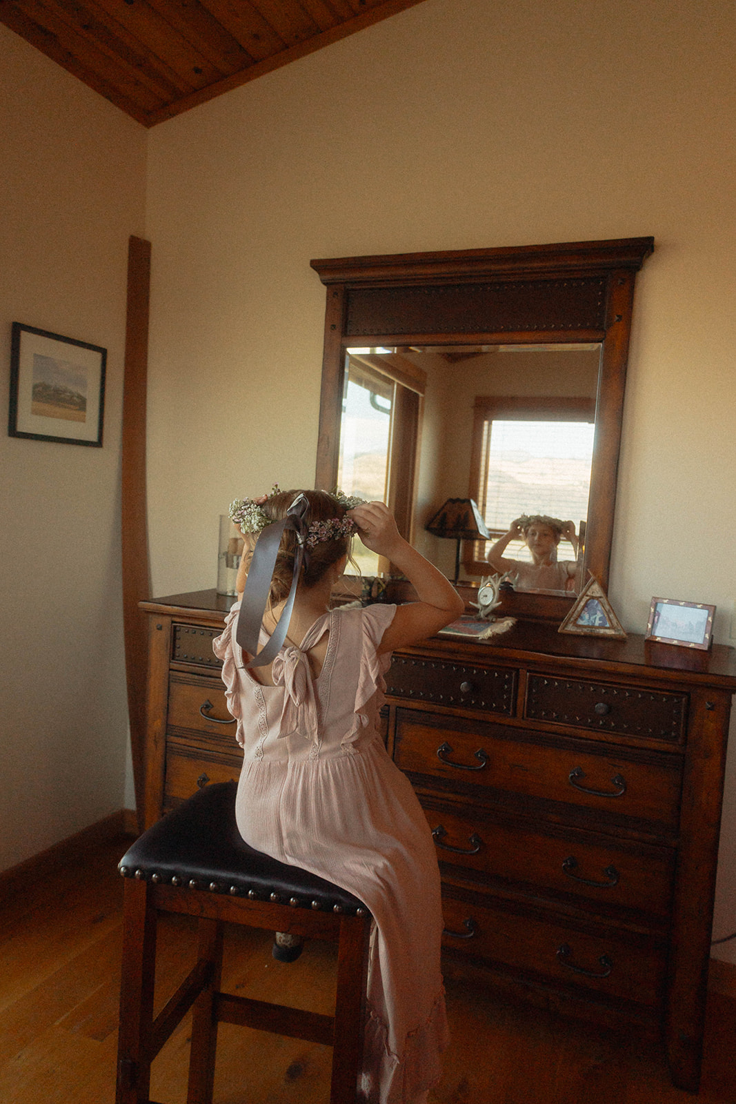 A person in a long dress sits on a stool in front of a wooden dresser with a mirror, adjusting their hair with both hands. The room has beige walls, a wooden floor, and a framed picture on the wall.