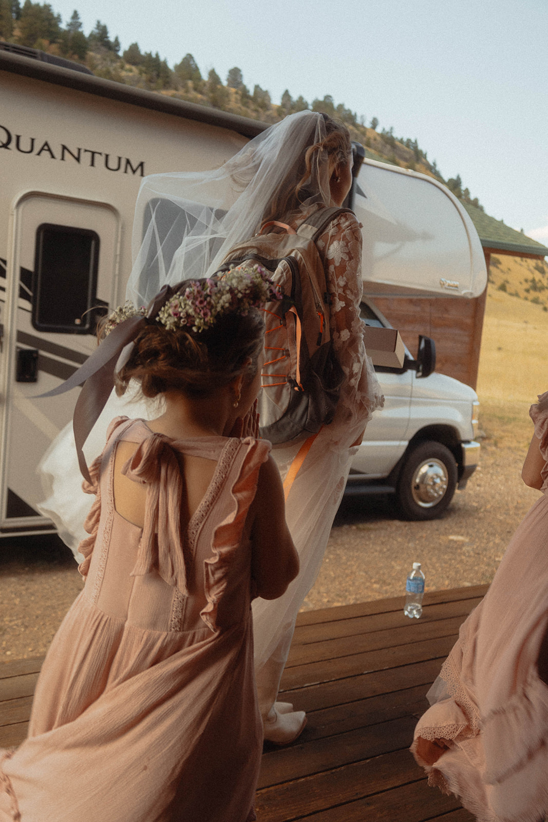 Two children in light pink dresses walk behind a woman in a floral dress and veil near a trailer. Dry, grassy hills are in the background.