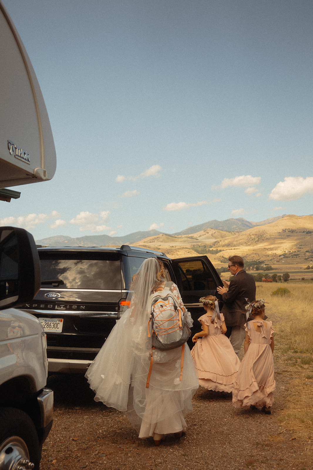 Two children in light pink dresses walk behind a woman in a floral dress and veil near a trailer. Dry, grassy hills are in the background.