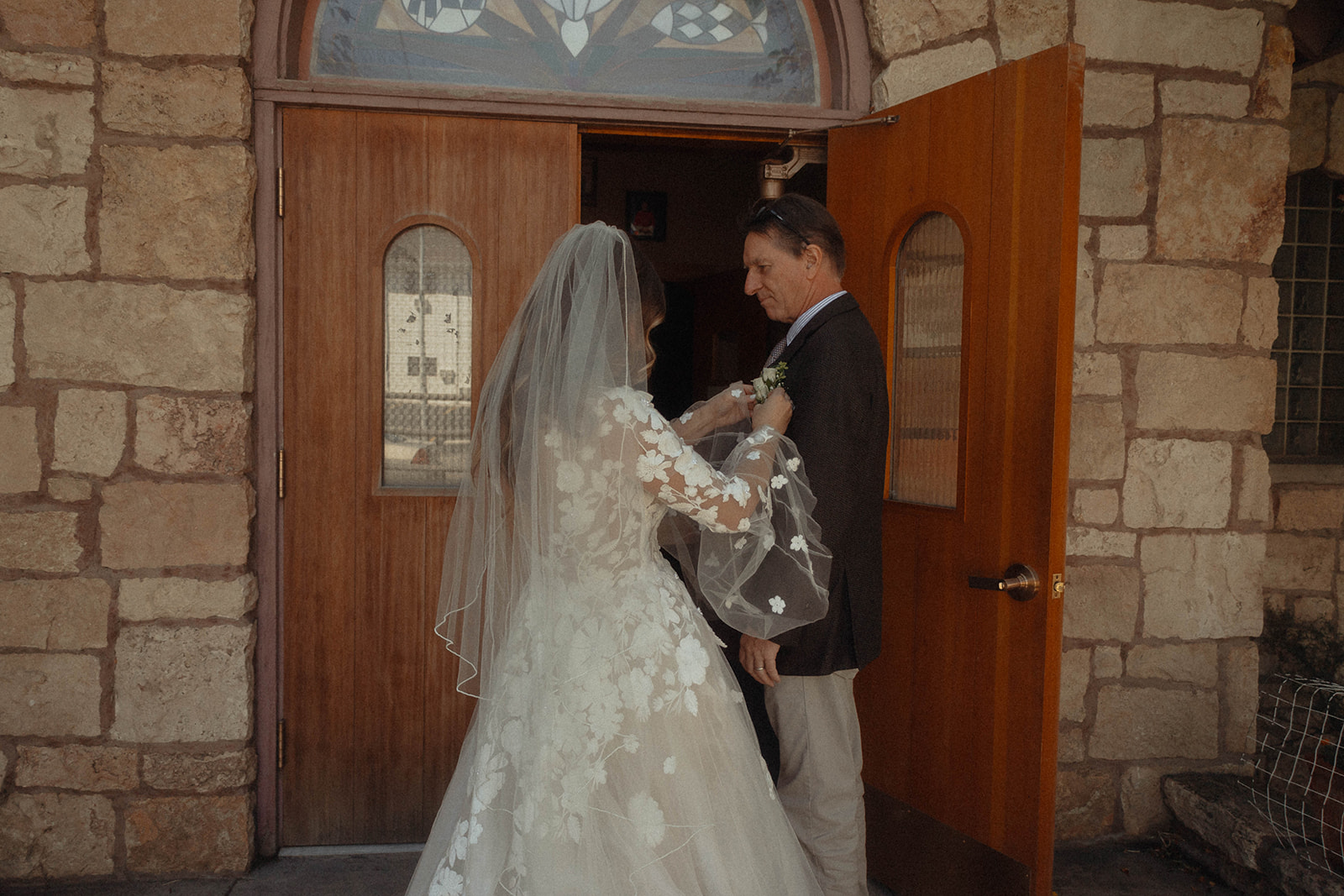 A bride and groom stand at the entrance of a stone building, with the bride adjusting the groom's boutonniere. The bride wears a lace-embroidered gown and veil, and the groom is in a dark suit.