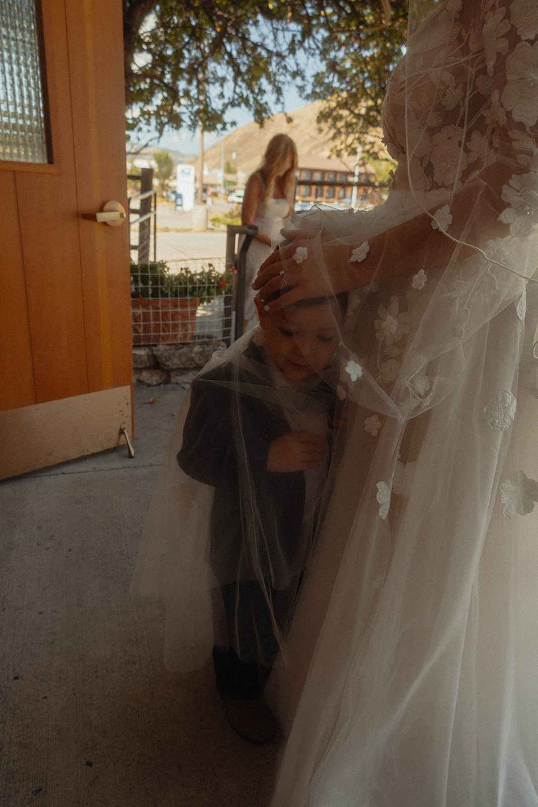 A child stands under a bride's veil, partially obscured by it. Another person is visible in the background outside. | children at your wedding