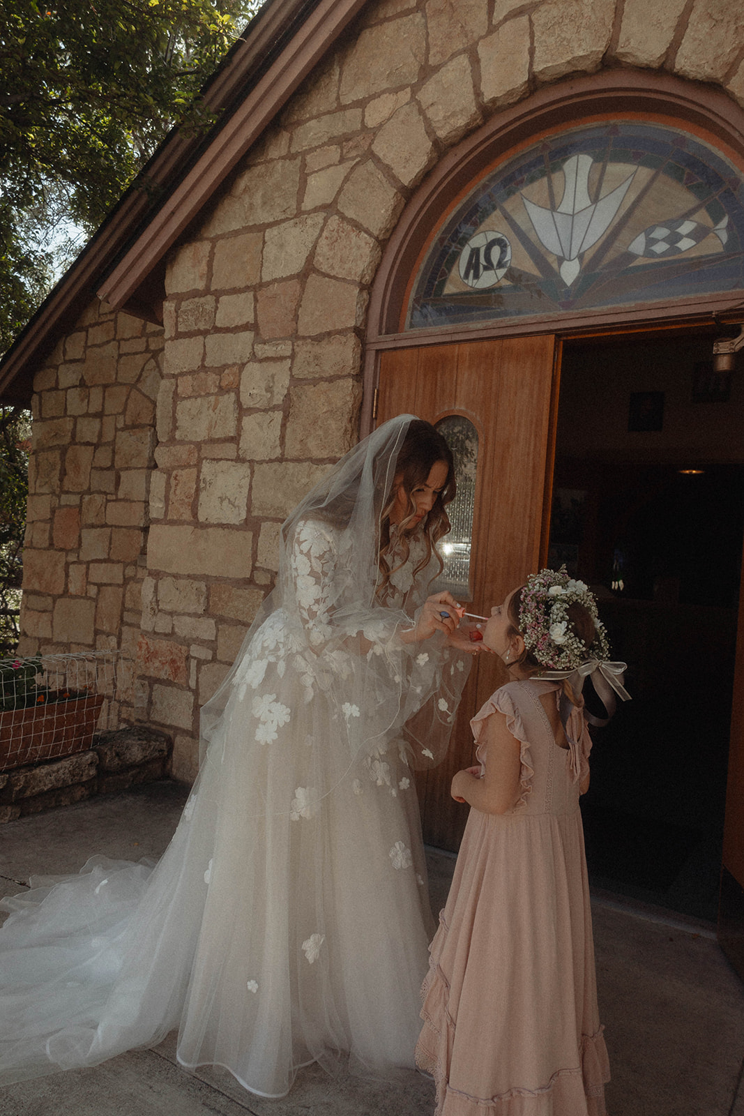 A woman in a white dress kneels on a pavement, smiling, with a child standing in front of her and a toddler sitting on her lap, both wearing dark suits | children at your wedding