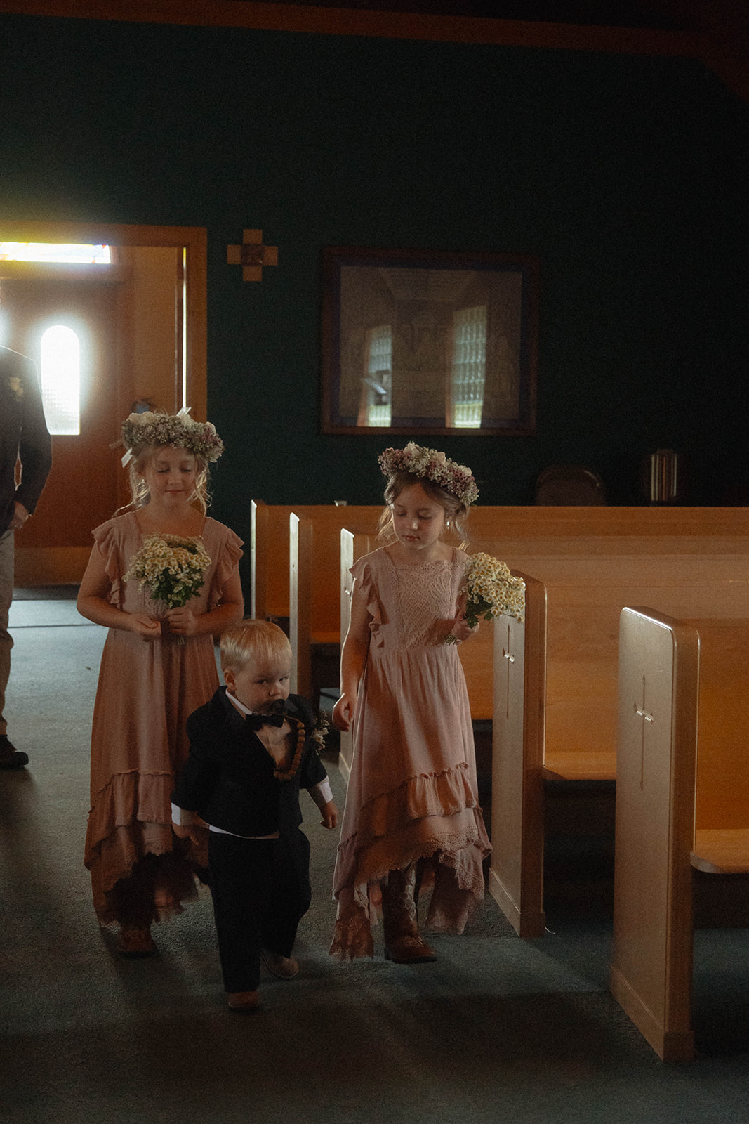 Three children, two girls in flower crowns and pink dresses holding bouquets, and a younger boy in a suit, walk down an aisle inside a dimly lit building | children at your wedding