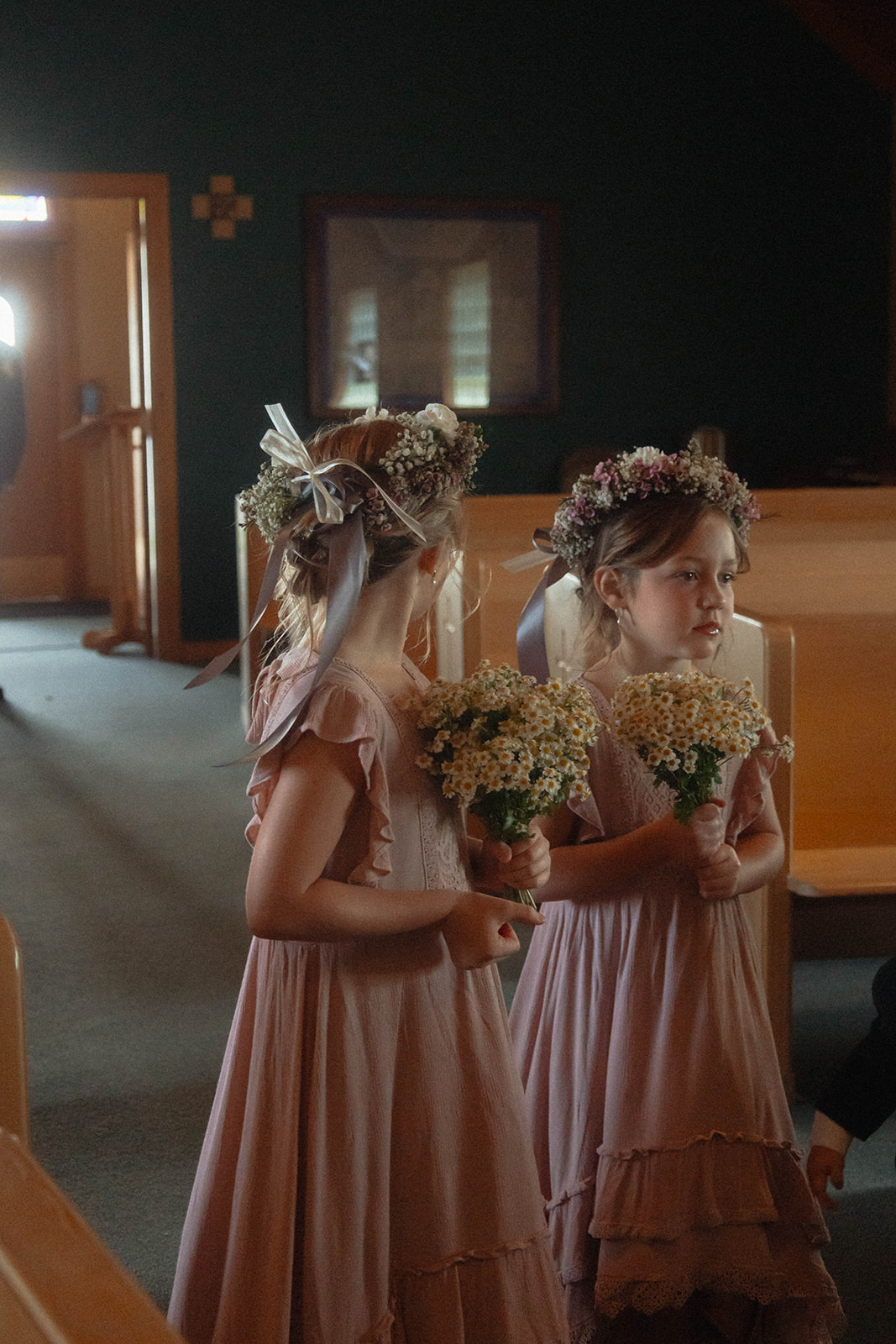 Three children, two girls in flower crowns and pink dresses holding bouquets, and a younger boy in a suit, walk down an aisle inside a dimly lit building | children at your wedding