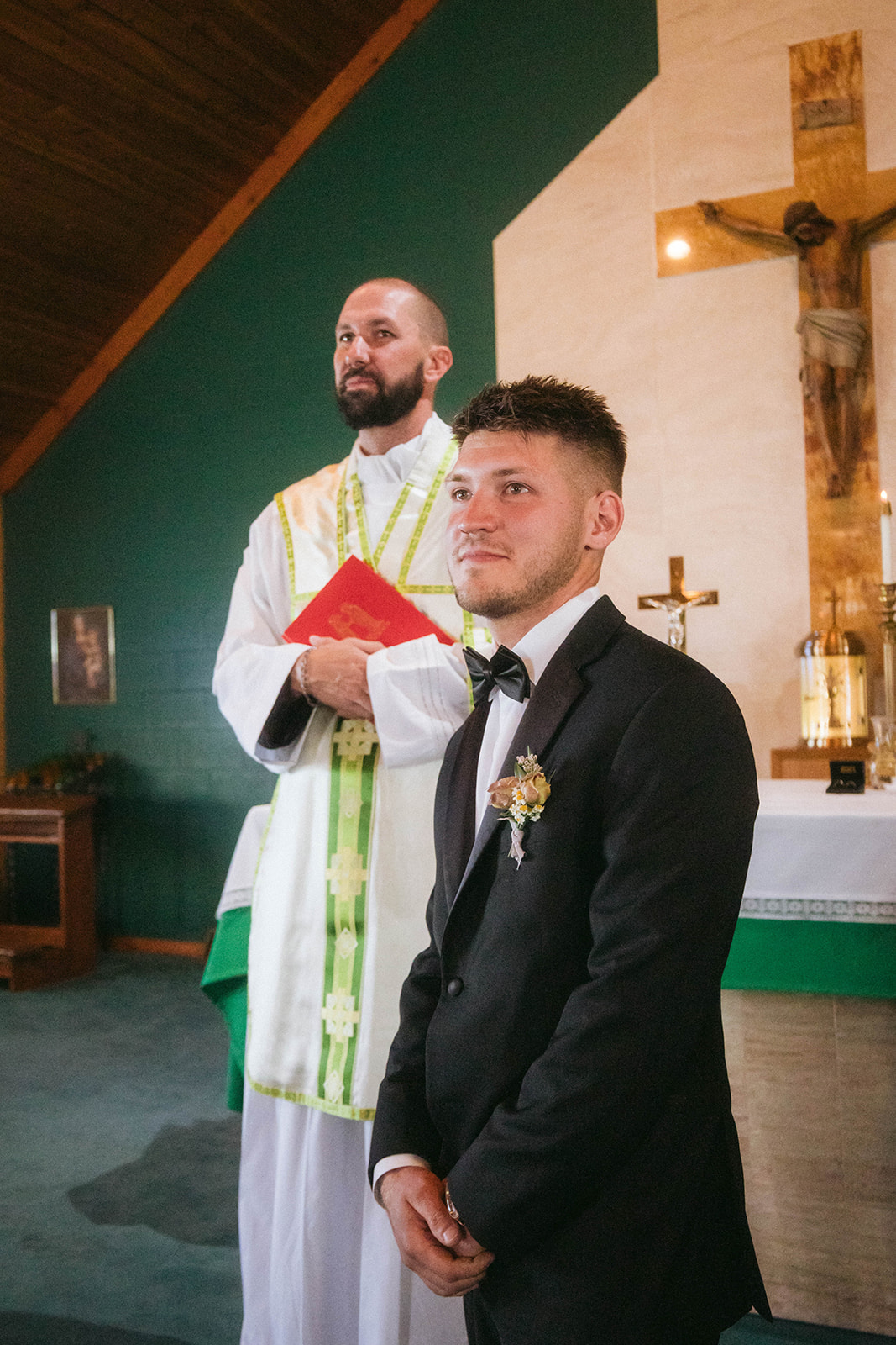 A groom in a black suit with a boutonniere stands beside a priest in white robes holding a red book inside a church