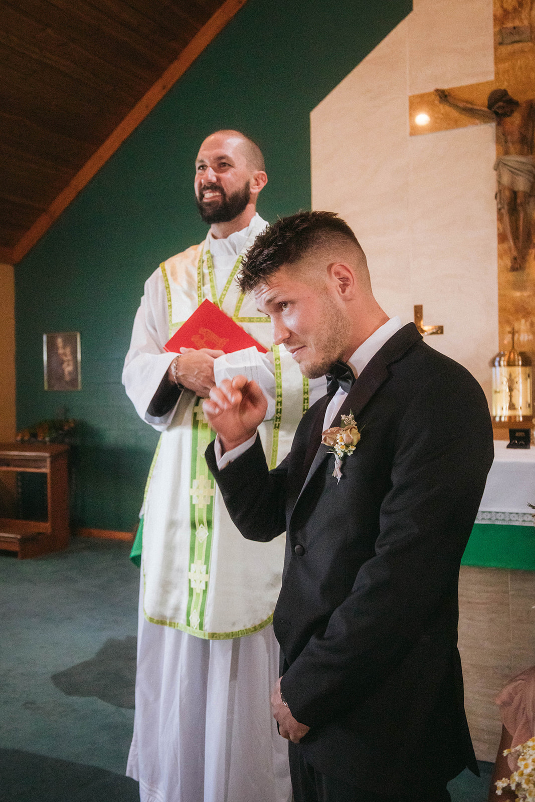 A groom in a black suit with a boutonniere stands beside a priest in white robes holding a red book inside a church 