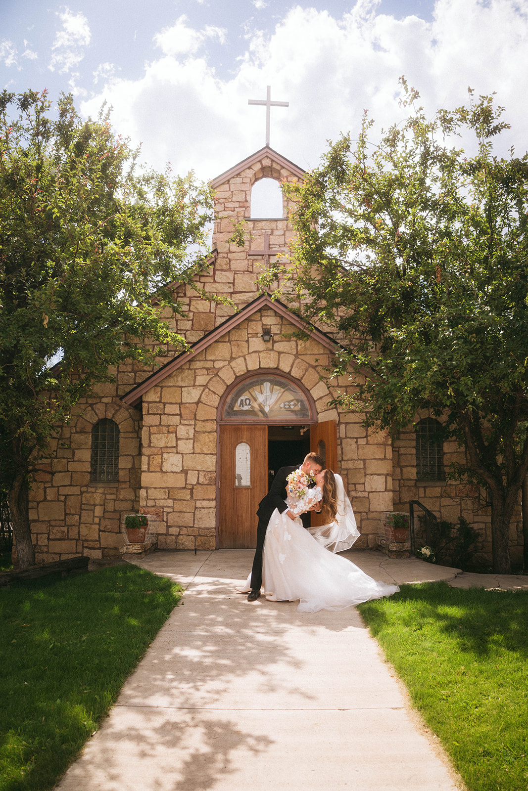 A couple in wedding attire stands in front of a stone church entrance, with trees flanking the building for a wedding in Gardiner, Montana