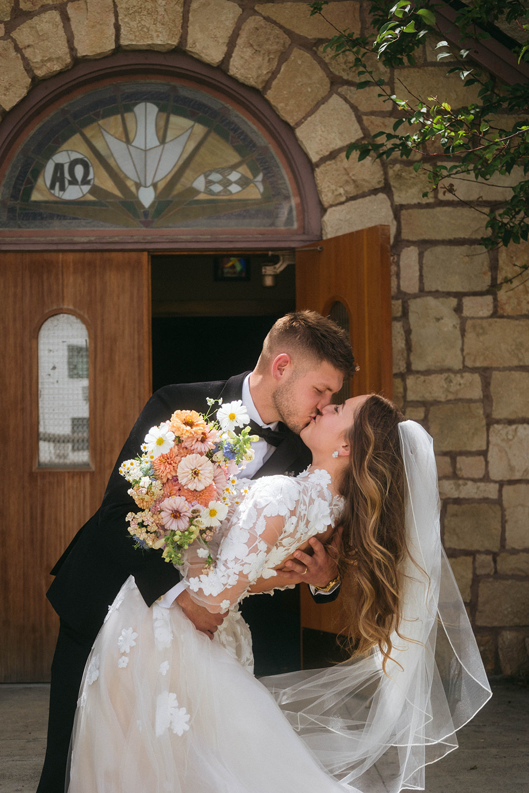 A couple in wedding attire stands in front of a stone church entrance, with trees flanking the building for a wedding in Gardiner, Montana