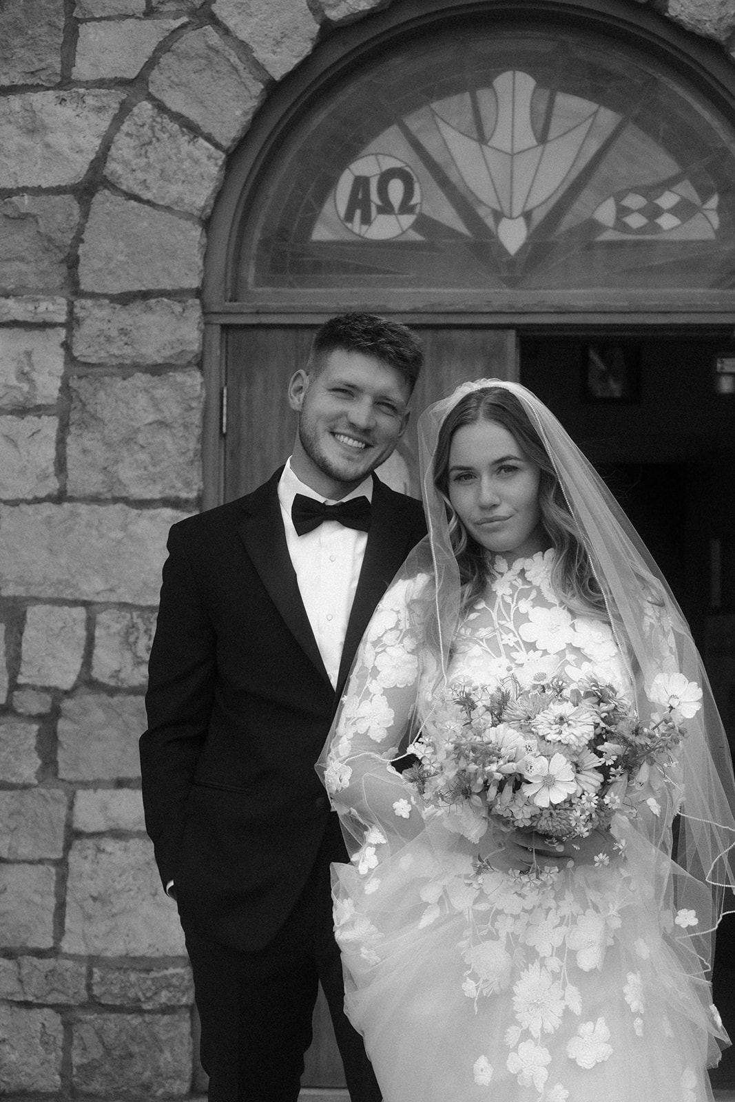 A couple in wedding attire stands in front of a stone church entrance, with trees flanking the building for a wedding in Gardiner, Montana