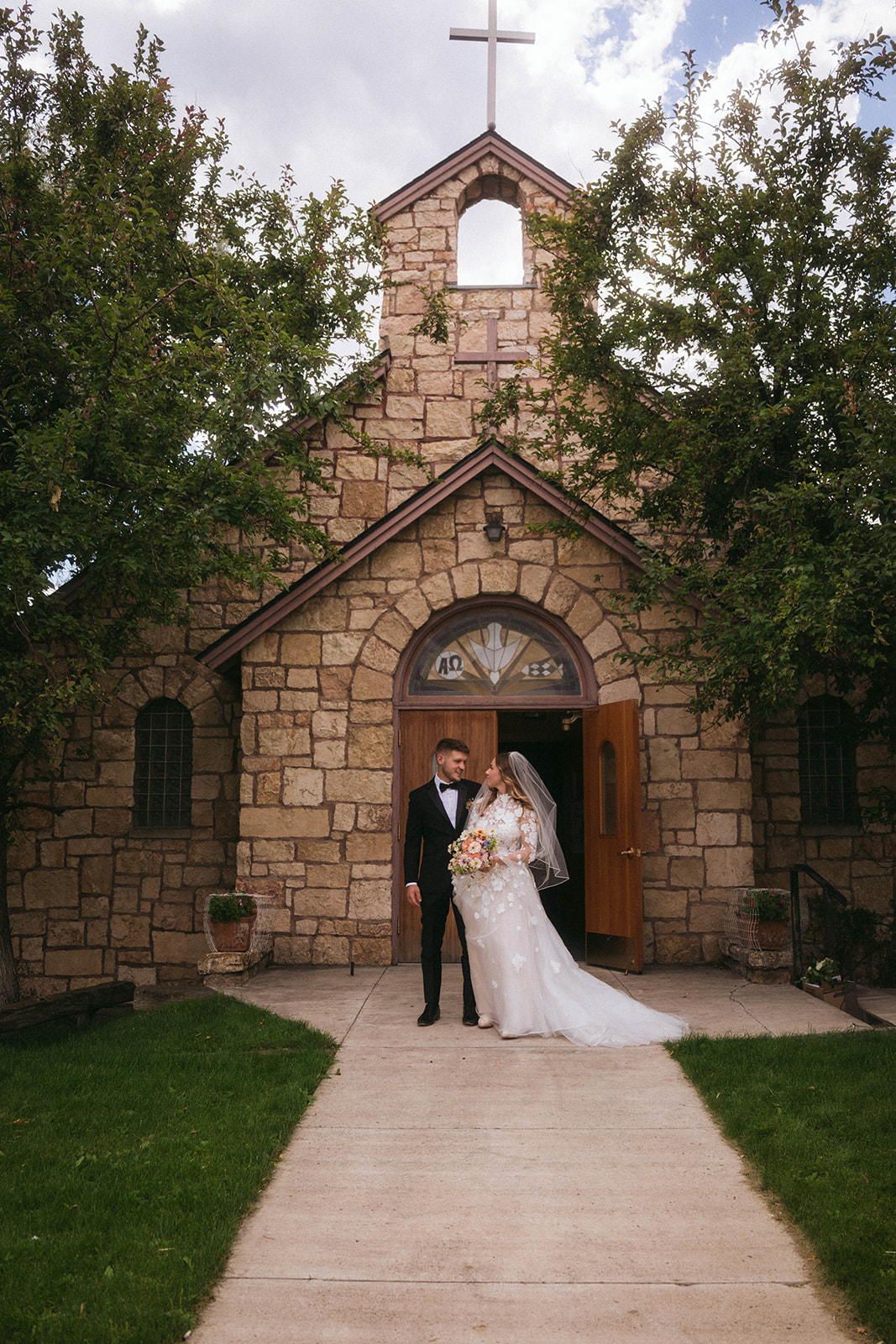 A couple in wedding attire stands in front of a stone church entrance, with trees flanking the building for a wedding in Gardiner, Montana