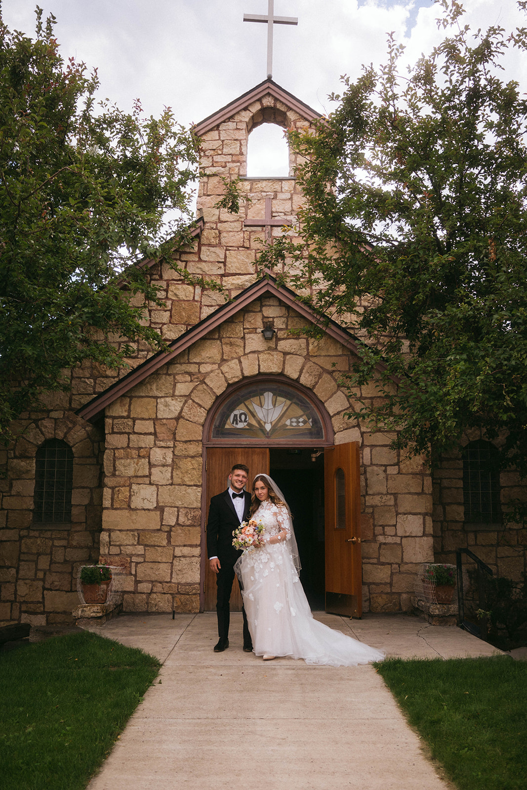 A couple in wedding attire stands in front of a stone church entrance, with trees flanking the building for a wedding in Gardiner, Montana